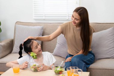 Neşeli bir anne ve kız besleyici yemeklerle kaynaşıyor, salatanın ve sağlıklı içeceklerin tadını çıkarırken parlak bir yuva ortamında kahkahalarını paylaşıyor..