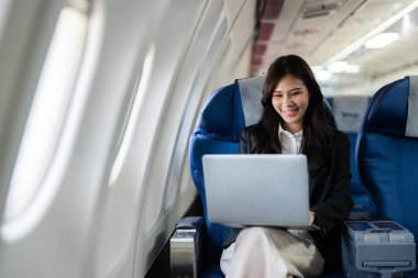 Uçakta otururken dizüstü bilgisayar kullanan odaklanmış bir iş kadını, seyahat ve uzaktan çalışmada verimliliği temsil ediyor.