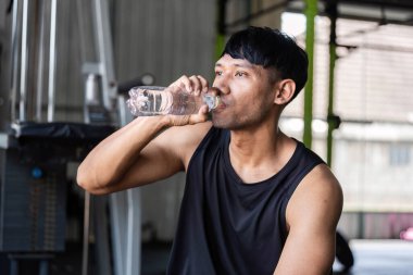 Spor salonunda dinlenirken bir adam serinletici bir su alır, vücut geliştirmede hidrasyonun önemini vurgular..