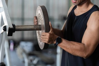 Genç bir adam ağırlık plakasını kaldırmaya hazırlanıyor. Spor salonunda spor yapmaya kararlı olduğunu gösteriyor..