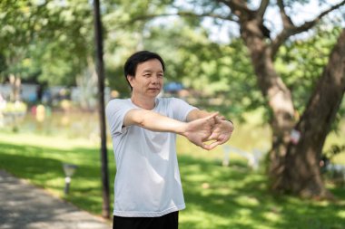 Yaşlı bir adam kollarını açık havada açar, sakin bir park atmosferinde farkındalık ve rahatlamaya odaklanır..