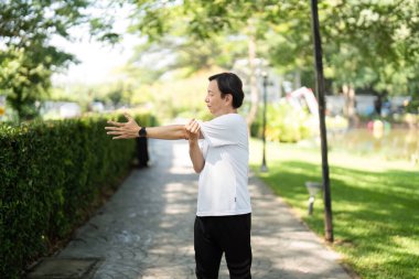 Yaşlı bir adam, sakin bir park ortamında esneklik ve sağlığın önemini vurgulamak için dışarıda kol esnetiyor..