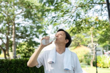 Bir adam parkta yürürken su içmek için biraz zaman ayırır. Sıhhatin sağlığa olan önemini vurgular..