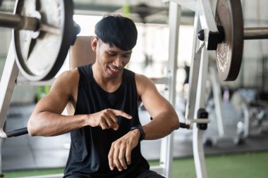 Genç bir adam spor salonunda otururken bileğindeki fitness izleyicisini kontrol ediyor. Teknolojinin modern fitness 'teki rolünü vurguluyor..