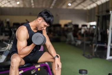 Bir erkek spor salonunda dambılı kaldırırken şekline odaklanır ve güç eğitimine olan bağlılığını vurgular..