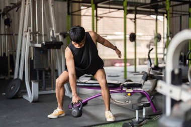 Kettlebell çalan genç bir adam spor salonunda ağırlık kaldırıyor, antrenman rutinini ve spora olan bağlılığını vurguluyor..