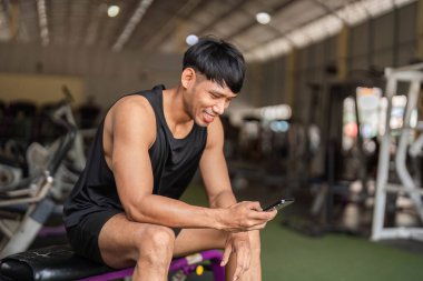 Sporcu genç bir adam akıllı telefonuna bakarken gülümser, spor salonunda mola verir. Fitness izleme ve motivasyon kavramı.