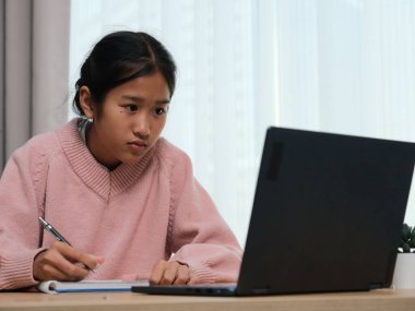 Masasında dizüstü bilgisayarı ve not defteri olan bir kız oturuyor. Pembe bir kazak giyiyor ve işine odaklanmış durumda.