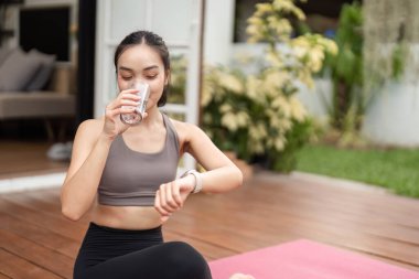 Genç bir kadın yoga pozunda oturur, su içer ve saatini kontrol eder, günlük rutininde sağlığını ve farkındalığını arttırır..