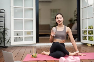 Genç bir kadın yoga pozunda oturur, suyu yudumlar ve etrafı sağlıklı yiyeceklerle çevrilidir. Evdeki sağlık ve zindeliği temsil eder..