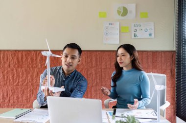 İki profesyonel ESG stratejilerini tartışırken, iş dünyasındaki sürdürülebilirliği vurgulayan bir rüzgar türbini modelini inceliyor.