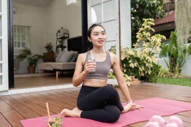 Yoga pozunda oturan, su içen ve sağlık rutininin bir parçası olarak ev ortamında susuzluğunu gideren bir kadın..