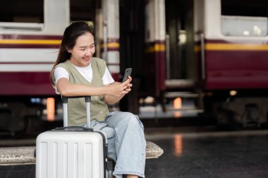 Tren istasyonunda oturan bir kadın, modern seyahat alışkanlıklarını yansıtan trenini beklerken telefonundaki güncellemeleri zevkle kontrol ediyor..