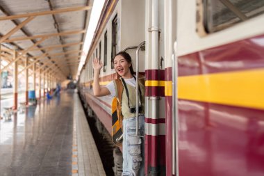 Yolculuğuna başlamak için heyecanlı olan bir gezgin, neşeli bir şekilde tren kapısından el sallıyor, canlı bir istasyonda macera ve keşfin özünü yakalıyor..