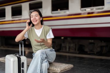 Neşeli bir gezgin, bir tren istasyonundaki telefon görüşmesi sırasında gülüyor. Seyahat etmenin ve sevdikleriyle iletişim kurmanın neşesini vurguluyor..