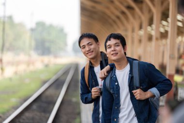İki arkadaş gülümser ve tren istasyonunda fotoğraf için poz verir. Sırt çantaları maceraya hazırdır. Sahne onların heyecanını ve yoldaşlığını yakalıyor..