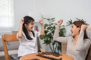 Bu görüntü, bir anne ve kızı zafer anında yakalıyor, eğlenceli ve canlı domino oyunlarını kutlamak için ellerini havaya kaldırıyor, neşe ve bağlantıyı simgeliyor..
