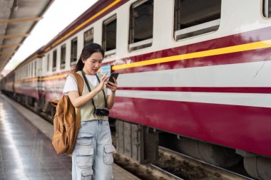 Genç bir kadın tren istasyonunda duruyor, bir sonraki yolculuğuna çıkmadan önce akıllı telefonuyla seyahat planlarını gözden geçiriyor..