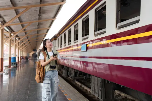 Yanında akıllı telefonuyla, genç bir kadın düşünceli bir şekilde tren istasyonuna bakıyor, yolculuğunun tadını çıkarıyor..