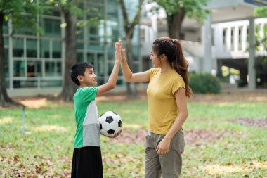 Genç bir çocuk, canlı bir park ortamında futbol antrenmanlarını ve takım çalışmalarını kutlayan bir kadına beşlik çakıyor..