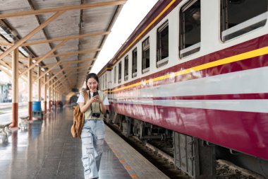 Tren istasyonunda telefonunu kullanan bir gezgin, bir sonraki macerasını beklerken heyecanla gülümsüyor..