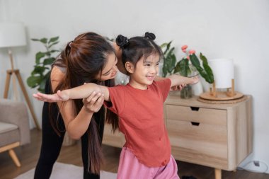 Bir anne, kızına destek olurken birlikte esneme egzersizleri yapar, hareketlerini teşvik eder ve neşeli bir ev ortamında iletişim kurar..