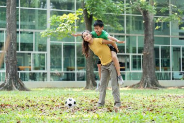 Yaratıcı bir an. Bir çocuk kollarını uçarak açar. Annesini sırtında taşırken, parkta oyun oynamanın zevkini ön plana çıkarır..