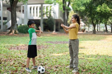 Genç bir çocuk ve bir kadın, futbol antrenmanından önce esneme ve takım çalışmasının önemini vurgulayarak parkta ısınma egzersizi yapıyorlar..