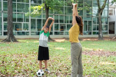 Hayat dolu bir park ortamında, bir kadın, yaklaşan futbol antrenmanları için eğlenceli bir ortam yaratarak, bir çocuğa esneme egzersizleri yaptırıyor..