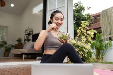 Yoga minderinde taze salata yerken gülümseyen genç bir kadın sağlıklı beslenme ve yaşam tarzını teşvik ediyor..