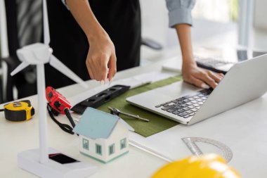 Bu sahnede, bir profesyonel dizüstü bilgisayar üzerinde çalışırken sürdürülebilir evler ve rüzgar türbinlerinin modellerini düzenler, teknolojiyi ekolojik tasarım ile harmanlar..