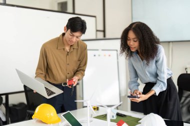 Gösterişli bir ofis ortamında, bir erkek ve bir kadın dizüstü bilgisayarlar ve minyatür modeller kullanarak sürdürülebilir enerji projeleri üzerinde işbirliği yapıyorlar, takım çalışması ve yenilik sergiliyorlar..