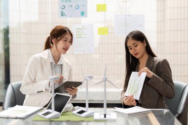 Gösterişli bir ofiste, iki kadın ESG stratejilerini tartışırken, sürdürülebilir çözümler ve daha iyi çevresel etki için veri güdümlü kararları vurgulayarak, çizelge ve modelleri gözden geçiriyorlar.