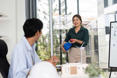 Genç bir kadın fikir sunarken, bir erkek not alır, takım çalışmasını ve sürdürülebilir tasarımda yeniliği ön plana çıkarır..