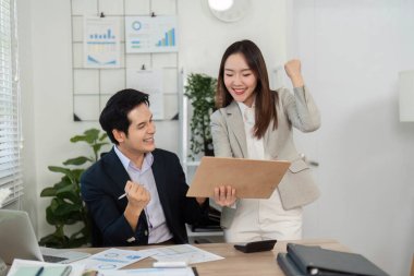 İki meslektaş heyecanlı bir şekilde finansal başarıları ve stratejileri parlak bir ofiste tartışıyor, takım çalışması ve işbirliği sergiliyor.