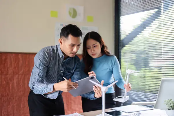 Bir ekip yenilenebilir enerji çözümlerini, bir rüzgar türbini modelini ve modern çalışma alanındaki tabletler üzerindeki dijital verileri analiz ederek tartışır..