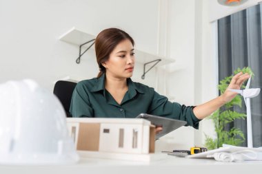 Bir kadın mimar, parlak bir ofiste proje doğruluğunu artırmak için tablet kullanırken sürdürülebilir bir modeli inceler..