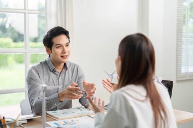 İki profesyonel ESG stratejilerini tartışırken, parlak bir ofiste rüzgar türbini modelleriyle çevriliyken sürdürülebilirlik girişimlerine odaklanıyorlar.