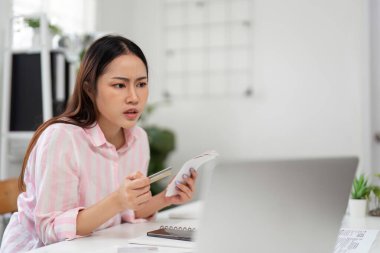 Endişeli bir kadın masasında oturuyor, faturalarını ve bilgisayar ekranını inceliyor, evden çalışırken online ödemelerini yönetme karmaşıklığıyla boğuşuyor..