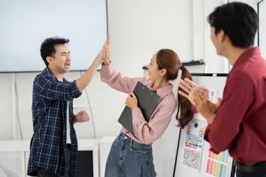 Canlı bir ofis ortamında, iki meslektaş coşkulu bir şekilde birbirlerine beşlik çakarken, üçüncü bir ekip üyesi de bu neşeli anı paylaşıyor, takım çalışması ve dostluk gösterisi yapıyor..