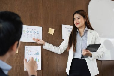 Modern bir ofis ortamında, bir kadın iş arkadaşlarına proje yönetim verilerini güvenle sunarak, işbirlikçi bir atmosferi teşvik eder..