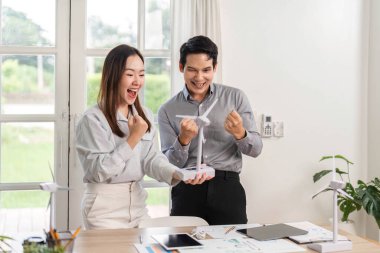 İki profesyonel, başarılı ESG projelerini parlak bir ofis alanında minyatür bir rüzgar türbini modeliyle sevinçle kutluyor..