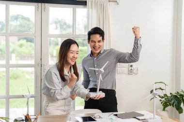 İki meslektaş, modern bir ofiste ESG ilkelerine ve yenilenebilir enerji çözümlerine duydukları heyecanı sergileyen bir rüzgar türbini modelini tartışıyor..