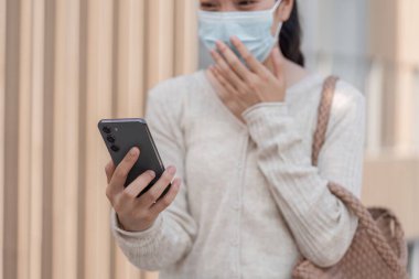 Maske takan bir kadın akıllı telefonunu tutuyor ve şehir merkezinde açık havada haberlere şaşırarak tepki veriyor..