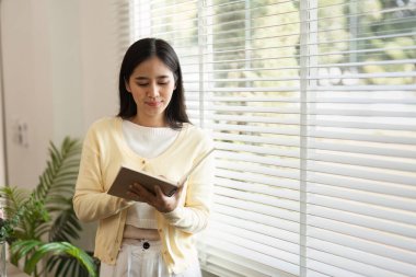 Genç bir kadın ev ofisinin penceresinin önünde duruyor, defterine odaklanmış, bir konsantrasyon ve yaratıcılık atmosferi yaratıyor..