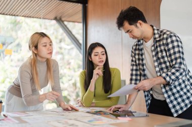 Çeşitli bir ekip tasarım materyallerini dikkatlice inceledi ve parlak bir ofis ortamında bir masanın etrafında kavramları tartıştı..