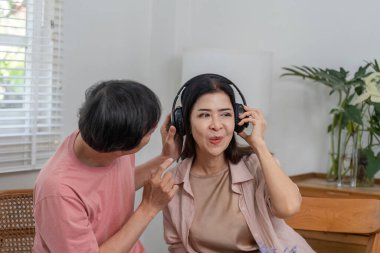 Yetişkin bir çift evde müzikle mutlu bir şekilde eğleniyor, bir eşi şakacı bir şekilde kulaklıklarını ayarlıyor, neşeli bir atmosfer yaratıyor..