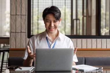 Genç bir işadamı, çağdaş bir ofis ortamında dizüstü bilgisayarındaki bir video görüşmesine katılırken gülümsüyor ve el kol hareketi yapıyor..