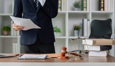 Bir avukat, elinde evrak tutarken ve dava detaylarını düşünürken hukuki belgeleri incelerken, çağdaş bir ofiste adalet terazileri ve kitaplar görülebilir..