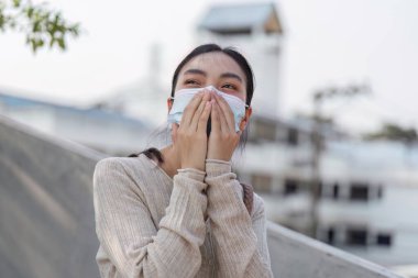 Maskeli heyecanlı bir kadın akıllı telefonuna bakarken geniş bir gülümseme sergiliyor, kirlenmiş kentsel ortamdaki pozitifliği yansıtıyor..